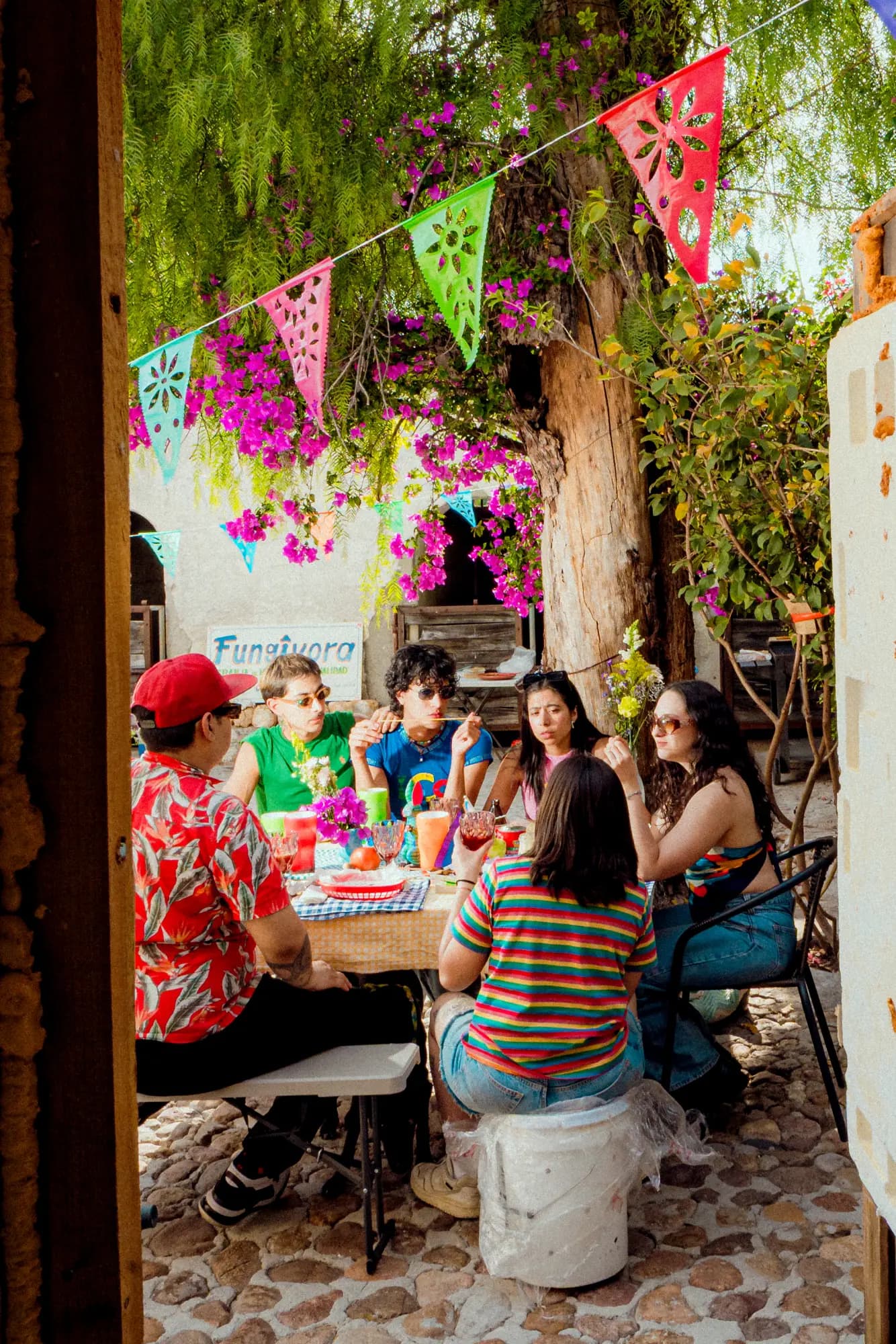 Fungívora, grupo de comensales compartiendo una comida al aire libre alrededor de una mesa en la granja, bajo papel picado de colores y buganvilias